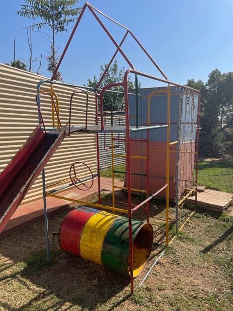 A brightly painted metal playground frame with a slide and an oil-drum tunnel on a sunlit lot in Soweto