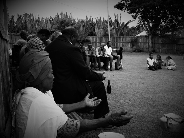 A black and white scene of a family gathering seated in a yard, children playing on the grass beyond